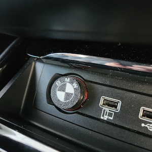 Op de afbeelding: Een zwarte en zilveren BMW auto sigarettenaansteker cover met het BMW logo. De ronde cover heeft een getextureerd oppervlak en een rode accent. Het is geïnstalleerd in een zwarte console met USB-poorten.