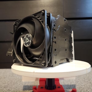 Op de afbeelding: Een zwarte CPU-koeler met een grote ventilator met het "ARCTIC"-logo. De koeler heeft een rechthoekige heatsink en is gemonteerd op een wit platform met een rode basis. De ventilatorbladen zijn zichtbaar en het apparaat heeft verschillende draden.