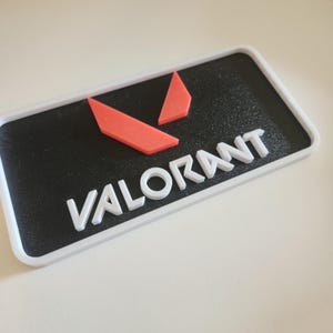 Puede incluir: Un emblema rectangular en blanco y negro con la palabra "VALORANT" en letras blancas. Encima del texto hay dos formas geométricas rojas. El emblema tiene un borde blanco.