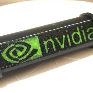 Könnte beinhalten: Schwarzes zylindrisches Objekt mit dem Wort "nvidia" in grünen Buchstaben. Das Nvidia-Logo ist ebenfalls grün. Das Objekt hat eine strukturierte Oberfläche und ein Gewindeende.