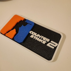 Op de afbeelding: Een rechthoekige patch met een witte rand met de tekst "COUNTER STRIKE 2" in het wit. De patch heeft een zwarte achtergrond met oranje en blauwe vormen aan de linkerkant.