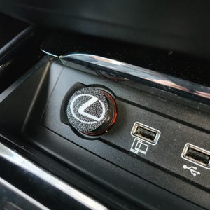 May include: A black and silver car cigarette lighter with the Lexus logo. The lighter is set in a black console with two USB ports. The lighter has a red accent ring.