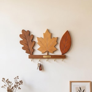 May include: A wall-mounted wooden key holder featuring three distinct leaf shapes in shades of brown, orange, and tan. The horizontal base includes five small brass-colored metal hooks, with a set of keys hanging from the second hook from the left.