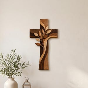 May include: A wall-mounted wooden cross featuring a layered, multi-toned brown design with an integrated carved branch and leaf motif that extends from the center, showcasing smooth, flowing curves and contrasting light and dark wood grain patterns.