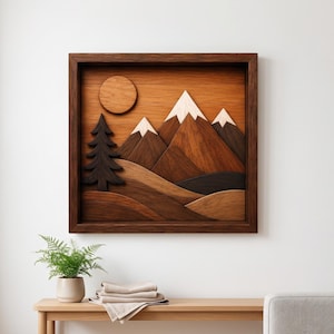 May include: A square wooden wall art piece featuring a layered landscape design. The artwork displays a dark brown pine tree, a circular sun, and mountain peaks with light-coloured tops, all set within a deep brown wooden frame.