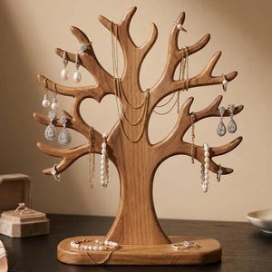 Puede incluir: Un expositor de joyas de madera de color marrón claro con forma de árbol y múltiples ramas para colgar collares, pendientes y pulseras. El soporte tiene un acabado liso y una base ovalada resistente, exhibiendo diversas piezas de joyería.