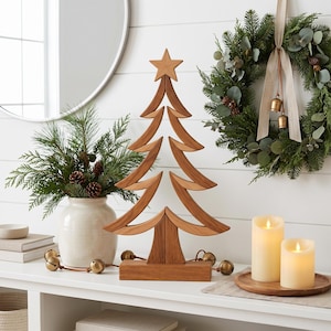 May include: A wooden Christmas tree decoration with a star topper, standing on a rectangular base. The tree is surrounded by a white ceramic vase with pine branches, a green wreath with a beige ribbon and bells, and two battery-operated candles.