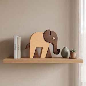 May include: A wooden elephant figurine crafted from light and dark wood tones, featuring a smooth, rounded body and a distinctively shaped ear and trunk. The elephant stands on two triangular dark wood supports, positioned next to two books.