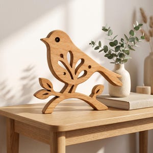 May include: A light brown wooden bird figurine perched on a branch. The bird features a cutout design on its wing with leaf-like shapes and a circular eye. The wood has a visible grain pattern, and the object stands upright on a flat surface.