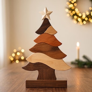 Op de afbeelding: Een decoratief houten kerstboombeeldje bestaande uit vijf gestapelde, golvende horizontale segmenten in verschillende tinten bruin en beige hout, bekroond met een lichtgekleurde houten ster op een rechthoekige houten voet.