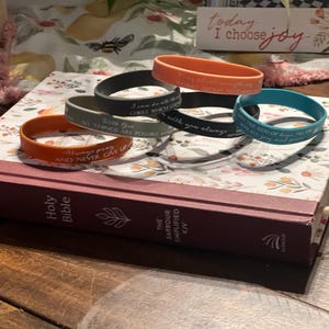 Könnte beinhalten: Eine Sammlung farbiger Silikonarmbänder mit inspirierenden Zitaten, die auf einer geschlossenen Heiligen Bibel liegen. Die Bänder sind orange, schwarz, türkis und grau. Die Bibel hat einen weinroten Einband und floral gemusterte Seiten.