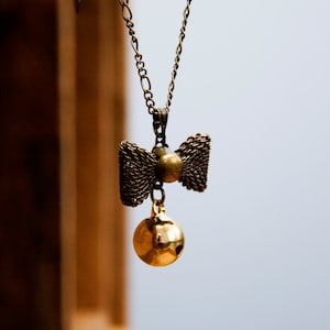 Könnte beinhalten: Eine goldene Kettenkette mit einem goldenen Schleifenanhänger und einem kleinen runden Anhänger. Die Schleife ist aus gewebtem Metall und der Anhänger ist glatt.