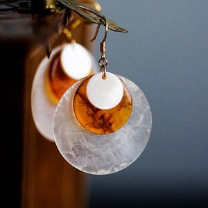 May include: A pair of gold hoop earrings with a white and amber layered design. The earrings are hanging from a wooden stand.