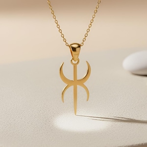 May include: A gold-colored pendant necklace featuring a stylized symbol. The pendant hangs from a delicate chain. The symbol has a vertical line with curved shapes on either side, and a small loop at the top. The necklace is displayed on a neutral-toned surface.