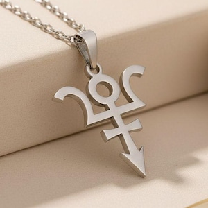 Könnte beinhalten: Eine silberfarbene Halskette mit Anhänger und einem stilisierten Symbol. Der Anhänger hat ein einzigartiges Design mit geschwungenen und eckigen Elementen, das an einer zarten Kette hängt. Die Halskette wird auf einer neutralen Oberfläche präsentiert, die ihren metallischen Glanz hervorhebt.