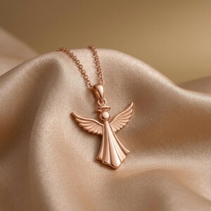 Puede incluir: Collar con colgante de &aacute;ngel en oro rosa. El colgante presenta un &aacute;ngel con alas extendidas y un vestido fluido. El collar se exhibe sobre una tela de sat&eacute;n. El colgante de &aacute;ngel mide aproximadamente 2,5 cm de altura.