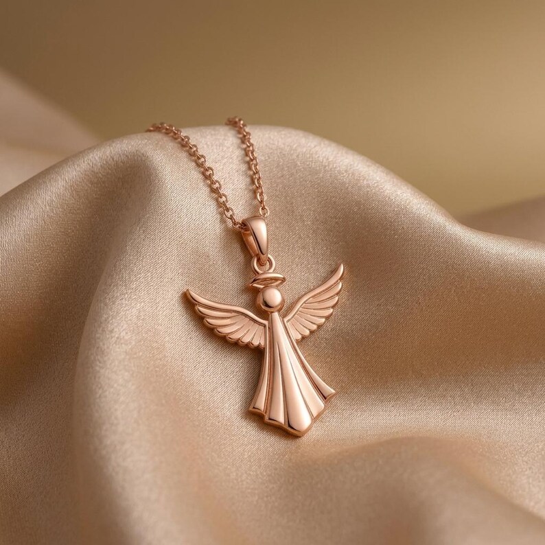 Puede incluir: Collar con colgante de &aacute;ngel en oro rosa. El colgante presenta un &aacute;ngel con alas extendidas y un vestido fluido. El collar se exhibe sobre una tela de sat&eacute;n. El colgante de &aacute;ngel mide aproximadamente 2,5 cm de altura.