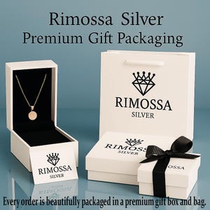 May include: A gold-colored pendant necklace displayed in an open white gift box with a black interior. The box is part of a set that includes a white gift bag, a small box tied with a black ribbon, and a card, all branded with the "Rimossa Silver" logo.