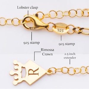 May include: Close-up of a gold-colored necklace with a lobster clasp and a 2.5-inch extender. The necklace features a crown-shaped pendant with the letter "R" and a "925" stamp. The chain is made of small, connected circles.