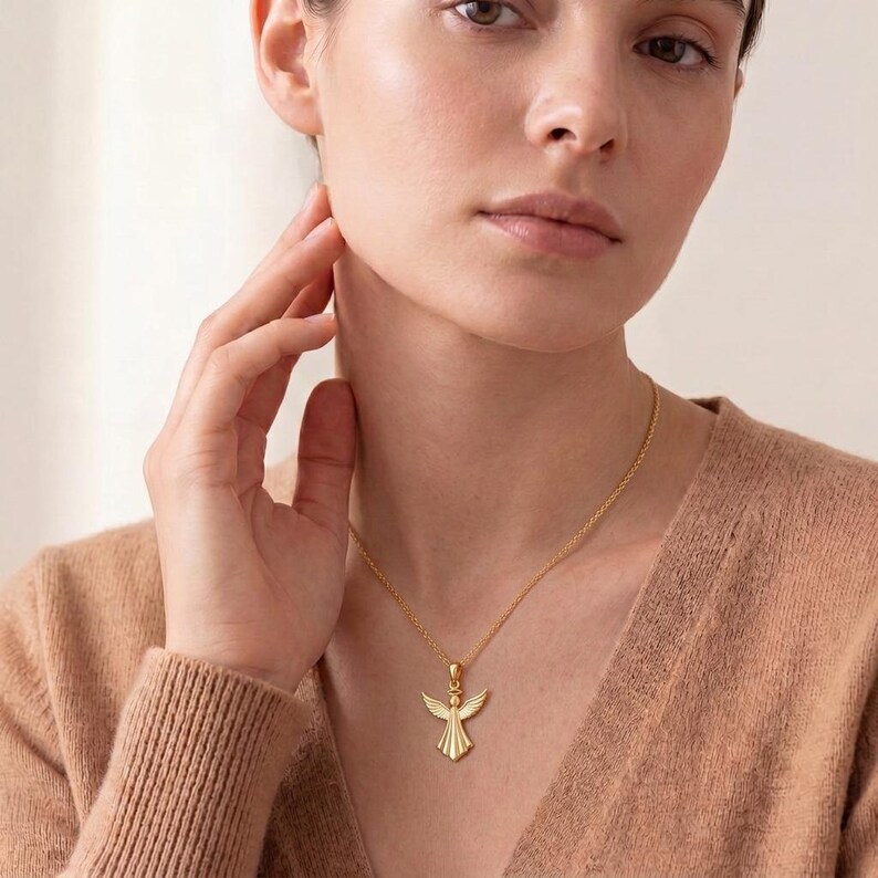 Puede incluir: Un collar con colgante de &aacute;ngel dorado. El colgante presenta un &aacute;ngel con las alas extendidas. El collar lo lleva una persona con un su&eacute;ter color beige. La cadena es delicada y el colgante est&aacute; detallado.