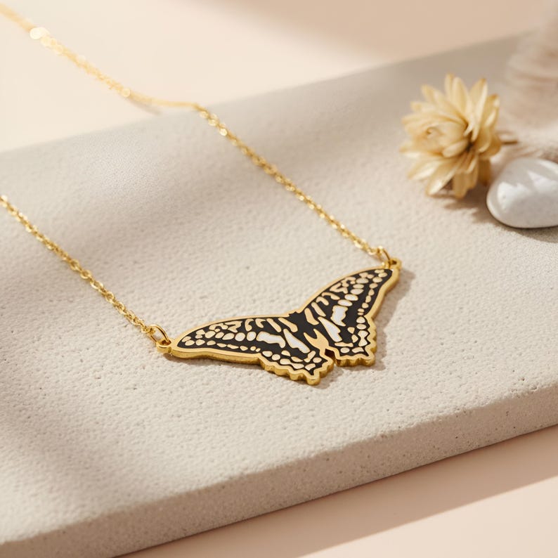 May include: A gold-colored butterfly pendant necklace. The butterfly design features black and white details. The necklace is displayed on a neutral-toned surface, with a dried flower and a small white stone in the background. The chain is delicate.