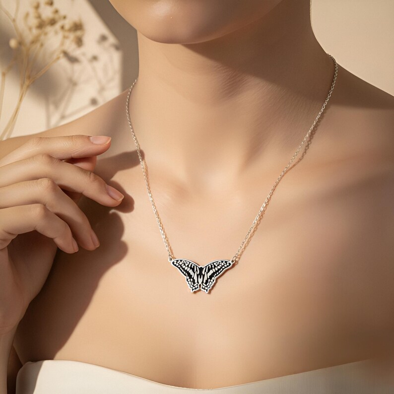 May include: A silver butterfly pendant necklace. The butterfly pendant features a black and white design. The necklace is worn around the neck. The background is a neutral color.