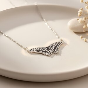May include: A silver necklace featuring a butterfly pendant. The butterfly pendant has a black and white pattern. The necklace is displayed on a white, round surface. The butterfly pendant is approximately 2 inches wide.