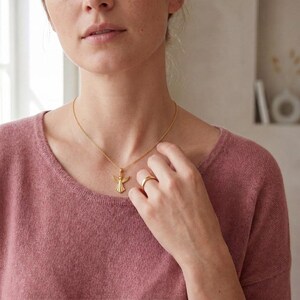 Puede incluir: Un collar dorado con un colgante de &aacute;ngel y un anillo dorado a juego. El colgante de &aacute;ngel tiene alas extendidas y un dise&ntilde;o detallado. El anillo es una banda simple. La persona lleva un su&eacute;ter rosa.