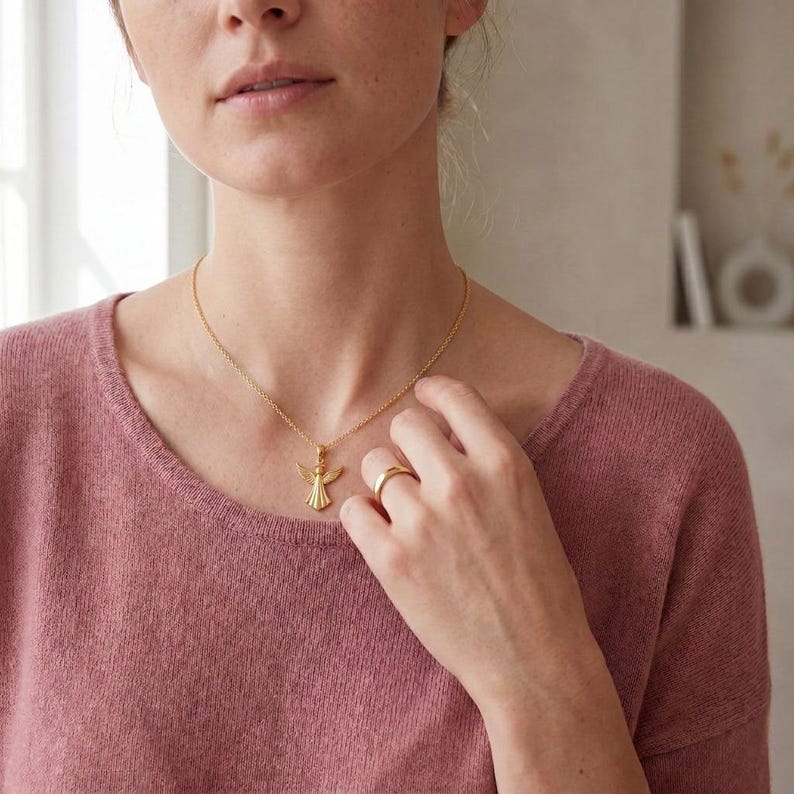 Puede incluir: Un collar dorado con un colgante de &aacute;ngel y un anillo dorado a juego. El colgante de &aacute;ngel tiene alas extendidas y un dise&ntilde;o detallado. El anillo es una banda simple. La persona lleva un su&eacute;ter rosa.