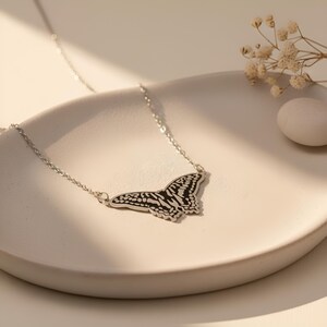 May include: A silver butterfly pendant necklace. The butterfly pendant features a black and white patterned design. The necklace is displayed on a white, curved surface, with dried flowers and a white stone in the background.