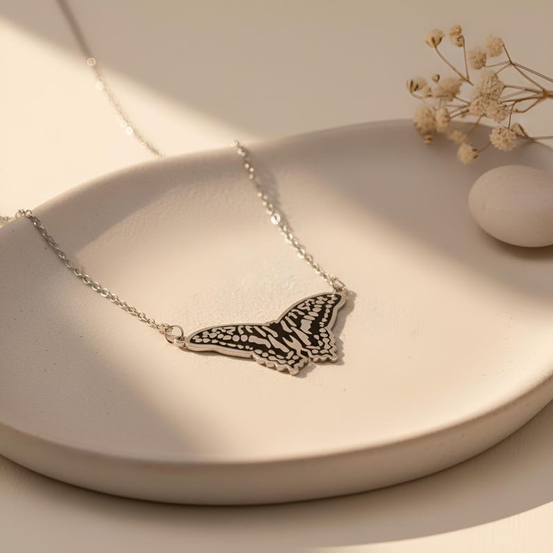 May include: A silver butterfly pendant necklace. The butterfly pendant features a black and white patterned design. The necklace is displayed on a white, curved surface, with dried flowers and a white stone in the background.