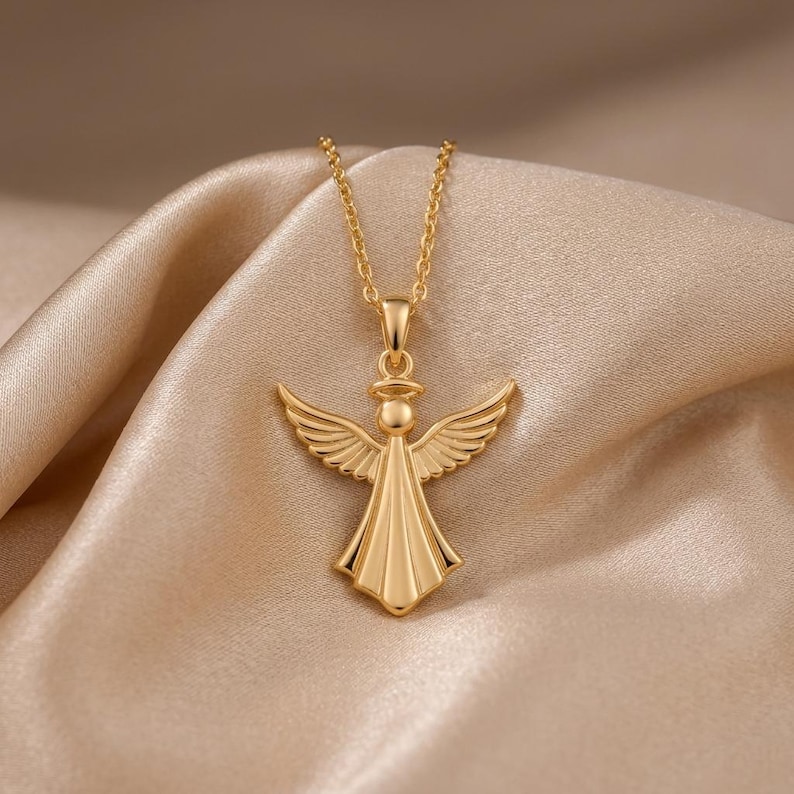 A polished angel pendant necklace with a delicate cable chain is displayed on a shimmering satin fabric. The angel pendant features outstretched wings, a halo, and a flowing gown, rendered in a smooth, reflective metallic finish. The necklace is available in both rose gold and gold tones, each subtly catching the light against the soft, champagne-colored background fabric. The chain drapes gracefully, highlighting the elegant design of the angel charm, which is the focal point of the image.