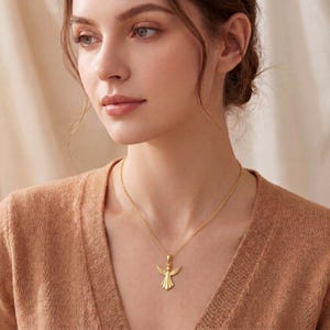 Puede incluir: Un collar con colgante de &aacute;ngel dorado. El colgante, con alas extendidas, cuelga de una fina cadena. El collar lo lleva una persona con un su&eacute;ter beige. Es una joya.