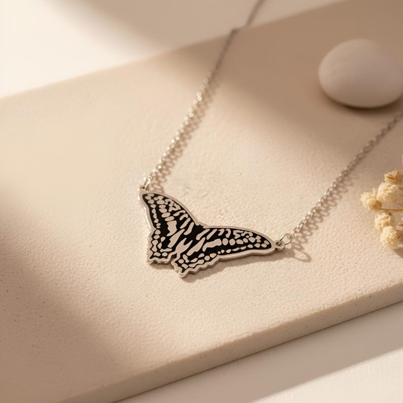 May include: A silver necklace featuring a butterfly pendant with a black and white patterned design. The butterfly pendant is the focal point, suspended from a delicate chain. The necklace is displayed on a neutral-toned surface.
