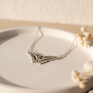 May include: A silver necklace featuring a butterfly pendant with black and white patterned wings. The necklace is displayed on a white, round plate, with dried flowers and a white object in the background.