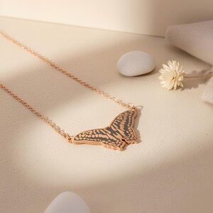 May include: A rose gold butterfly pendant necklace. The butterfly pendant has a detailed design with dark gray accents. The necklace is displayed on a light beige surface with a small white stone and a dried flower.