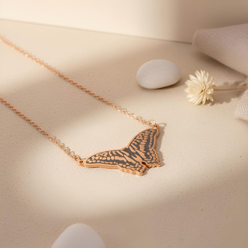 May include: A rose gold butterfly pendant necklace. The butterfly pendant has a detailed design with dark gray accents. The necklace is displayed on a light beige surface with a small white stone and a dried flower.