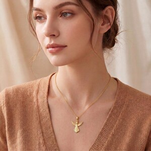A polished angel pendant necklace with a delicate cable chain is displayed on a shimmering satin fabric. The angel pendant features outstretched wings, a halo, and a flowing gown, rendered in a smooth, reflective metallic finish. The necklace is available in both rose gold and gold tones, each subtly catching the light against the soft, champagne-colored background fabric. The chain drapes gracefully, highlighting the elegant design of the angel charm, which is the focal point of the image.