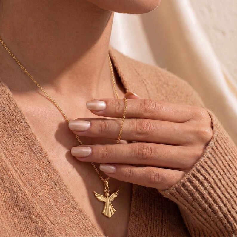 Puede incluir: Un collar con colgante de &aacute;ngel dorado. El colgante presenta un &aacute;ngel con las alas extendidas. El collar lo lleva una persona con un su&eacute;ter color beige. El collar es una joya delicada.