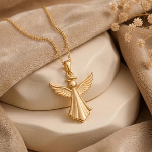 Puede incluir: Un collar con colgante de &aacute;ngel dorado. El colgante presenta un &aacute;ngel con alas extendidas y un halo. El collar se exhibe sobre una superficie beige con una tela suave y drapeada y ramitas de flores secas.
