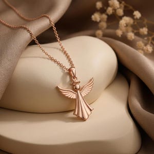 Puede incluir: Collar con colgante de &aacute;ngel en oro rosa. El colgante presenta un &aacute;ngel con alas extendidas y un vestido fluido. La cadena del collar es delicada y complementa el dise&ntilde;o del colgante. La joya se exhibe sobre un fondo neutro.