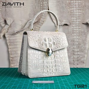 May include: A white crocodile-effect handbag with a top handle and a flap closure. The bag has a decorative clasp with black, white, and green details. The brand name "DAVITH" is visible at the top left. The product code "TG21" is at the bottom right.