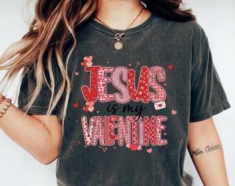May include: Dark gray t-shirt with the text "Jesus is my Valentine" in red and pink lettering. The design includes hearts and an envelope. The shirt is paired with blue jeans and white sneakers.
