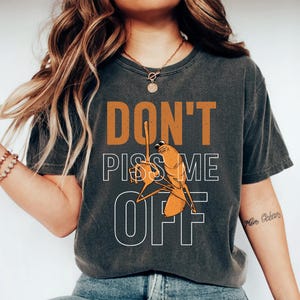 May include: Dark gray t-shirt with the text "DON'T PISS ME OFF" in orange and white. The shirt features a cartoon illustration of a bug with a raised middle finger. The person is wearing blue jeans and white sneakers.