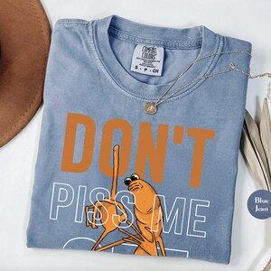 May include: Blue jean colored t-shirt with the words "DON'T PISS ME" in orange and white. The graphic features an orange shrimp with a raised middle finger. A gold necklace is draped across the shirt.