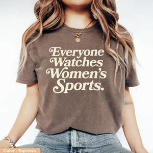 May include: Espresso-colored t-shirt with the text "Everyone Watches Women's Sports" in a cream-colored, vintage-style font. The shirt is a short-sleeved crew neck.