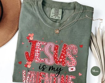 May include: A moss green t-shirt with the words "JESUS is my VALENTINE" in red and pink lettering. The design includes hearts and a key. A silver necklace with a pendant is on the shirt.
