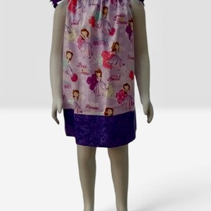 Puede incluir: Un vestido sin mangas con fondo morado claro y un patrón repetitivo de princesas de dibujos animados, flores y textos que incluyen "Hero", "Brave", "Adventures" y "Amulet". El vestido tiene lazos morados oscuros en los hombros y un dobladillo morado oscuro.
