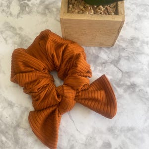 May include: A burnt orange hair scrunchie made of a ribbed fabric. The scrunchie features a knotted bow detail with two fabric tails extending from the center. The material has a distinct vertical texture throughout the entire accessory.
