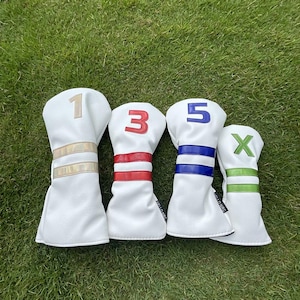 May include: Four white golf club head covers with colored accents. The covers are numbered 1, 3, 5, and X. Each cover has a different color scheme: gold, red, blue, and green. The covers are arranged on a green grassy surface.